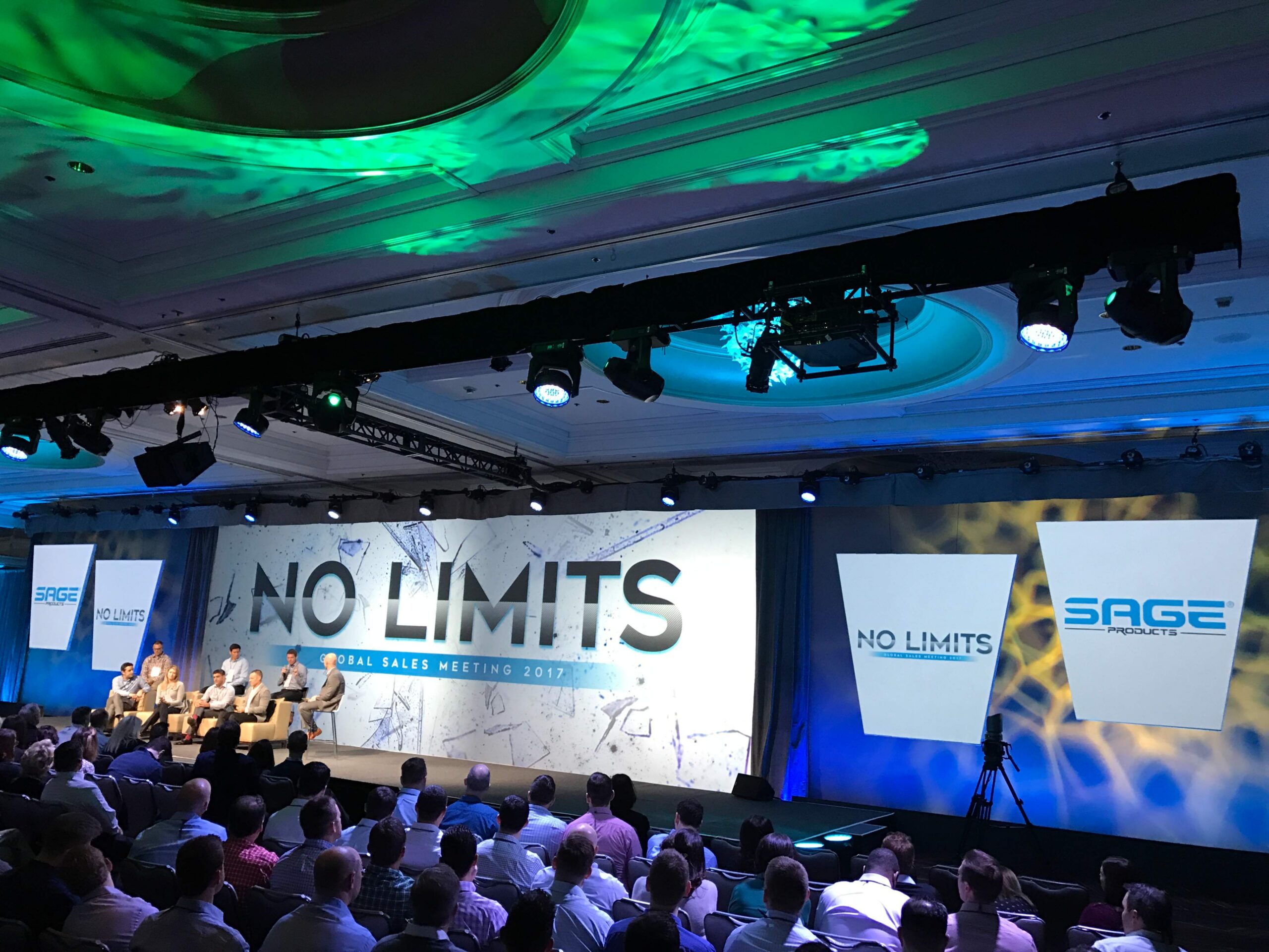 A group of people in a panel discussion at a corporate event are sitting on stage in front of a large screen.